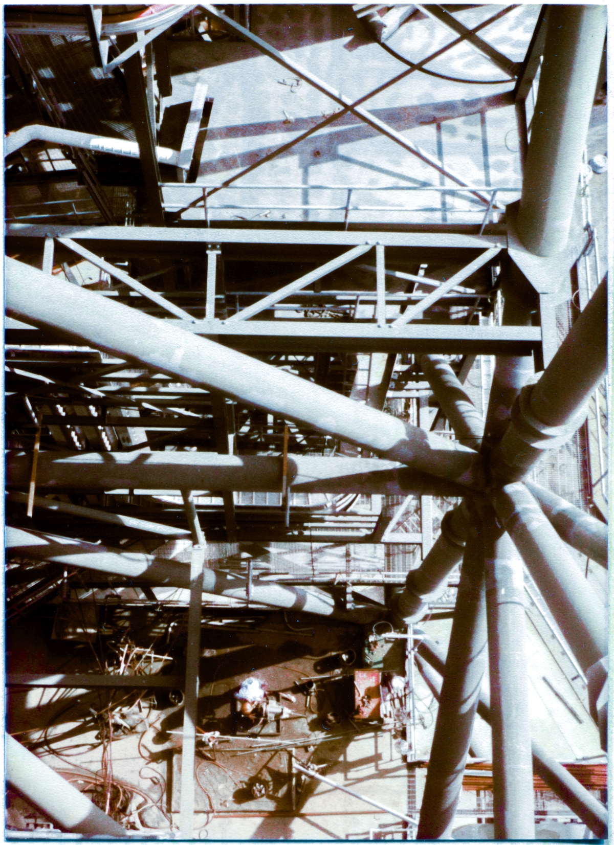 Image 022. You are standing with your boots on the steel bar grating at elevation 212’-7”, leaning out as far past the perimeter handrail as you dare, eighty feet above the Main Floor Level of the Rotating Service Structure at Space Shuttle Launch Complex 39-B, Kennedy Space Center Florida, looking vertically down and across toward that main floor at elevation 135’-7”, where a worker in an orange hardhat is bent over his equipment, plying his trade. The heavy steel pipes of the Primary Framing structure of the RSS, along with a bewildering array of Secondary Framing steel, and Miscellaneous Metals steel, makes for an uncanny visual ambiance that is simultaneously appealing and confusing. Your point of view is over 200 feet above the bottom of the Flame Trench at the launch pad, a tiny sliver of which is barely visible, shrouded in deep shadow, at the far top right margin of this image, immediately to the right of a massive structural pipe. Photo by James MacLaren.